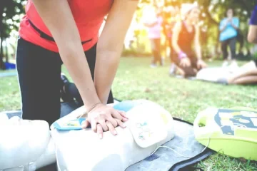 Person practicing CPR on a mannequin outdoors with an AED training device nearby, during a first aid training session.