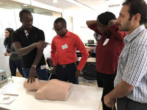 A group of people stand around a medical training mannequin, with one person demonstrating a procedure in a classroom setting.