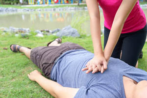 Person performing CPR chest compressions on an unresponsive individual lying on grass outdoors near a pond.