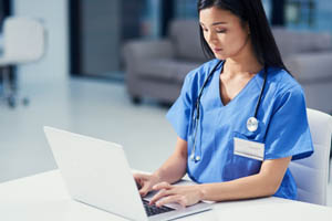 Female healthcare worker in blue scrubs using a laptop at a desk in a medical or hospital office setting.