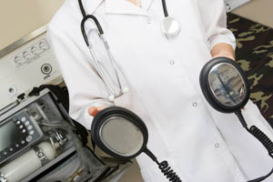 Doctor in a white coat holding defibrillator paddles, with a stethoscope around their neck and medical equipment in the background.