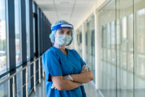Healthcare worker in blue scrubs, face shield, mask, and gloves standing in a hospital corridor with arms crossed.