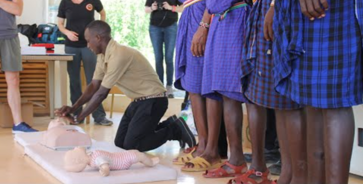 A man demonstrates CPR techniques on a mannequin while a group of people watch, some in blue plaid garments and sandals, during an indoor training session.