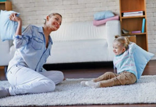 Woman and young child having a playful pillow fight on a carpeted living room floor with a couch and shelves in the background.