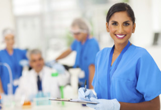 Smiling nurse in blue scrubs holding a clipboard, with medical team working in the background in a hospital or clinic setting.
