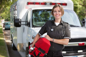 Female paramedic in uniform holding a red medical bag stands in front of an ambulance, smiling outdoors on a sunny day.