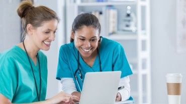 Two smiling female healthcare professionals in scrubs looking at a laptop together in a medical office, with a coffee cup on the desk nearby.