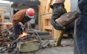 Worker holding a sledgehammer in the foreground with another worker in safety gear operating machinery in the background.