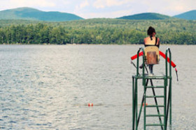 Lifeguard sitting on a high chair overlooking a calm lake with forested hills in the background.