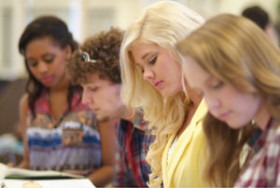 Four students sitting in a row, focused on reading or writing, in a classroom or study environment.
