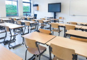 Empty classroom with rows of wooden desks and chairs, large windows, a digital screen, and a whiteboard at the front.