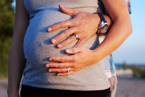 Close-up of a pregnant woman in a gray shirt with two hands, one male and one female, gently resting on her baby bump outdoors.