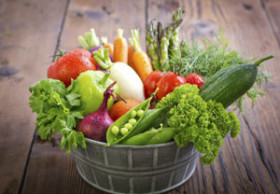 A metal bowl filled with fresh assorted vegetables, including tomatoes, carrots, onions, lettuce, cucumber, peas, asparagus, and parsley, on a rustic wooden table.