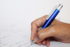 Hand holding a blue pen while writing on white paper with cursive text.