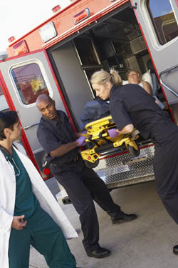 Paramedics unload a stretcher from an ambulance as a patient lies inside, while a doctor stands nearby.