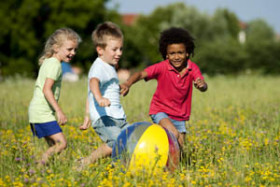 Three young children playing with a colorful beach ball in a grassy field filled with yellow wildflowers on a sunny day.