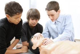Three boys practice CPR techniques on a medical training mannequin, with one demonstrating mouth-to-mouth resuscitation while the others observe closely.