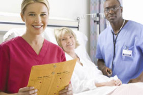 Smiling nurse holding a chart beside a female patient lying in a hospital bed, with a doctor standing nearby.