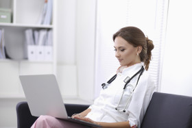 Female doctor with a stethoscope sitting on a couch and working on a laptop in a bright medical office.