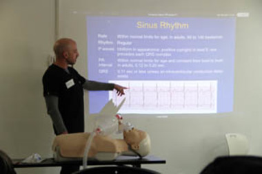 Instructor pointing at a sinus rhythm presentation slide while standing beside a CPR training mannequin in a classroom setting.