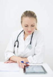 Female doctor in white coat with stethoscope reviews documents and tablet at a desk.