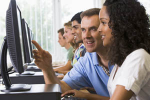 Group of adults sitting in a row at computers, working together and smiling in a bright computer lab or classroom.