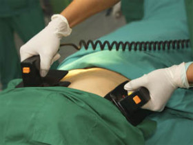 Medical professional using a defibrillator on a patient wearing green surgical drapes during an emergency procedure.
