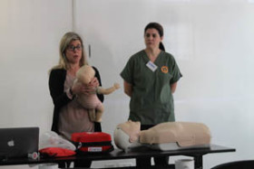 Two women demonstrate CPR techniques with infant and adult mannequins in a classroom setting with training equipment on the table.