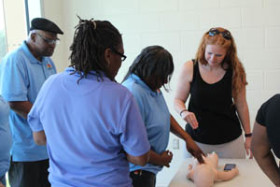 A group of adults gathered around a table practicing infant CPR techniques on a training mannequin.