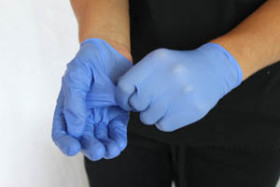Person wearing blue disposable gloves, using one gloved hand to remove the glove from the other hand against a plain background.