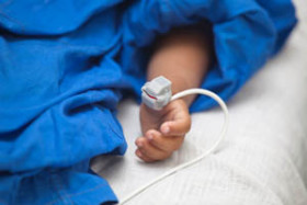 A child’s hand with a pulse oximeter sensor attached, lying on a hospital bed covered by a blue blanket.