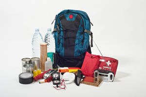 Emergency preparedness kit including a blue backpack, water bottles, canned food, flashlight, radio, first aid kit, batteries, tape, mask, gloves, and other survival supplies.