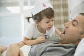 Young girl wearing a nurse hat pretends to examine a man's open mouth as he lies on a bed, engaging in playful role-play.