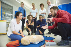 Instructor teaching CPR to a group of adults using a training mannequin and defibrillator in a classroom setting.