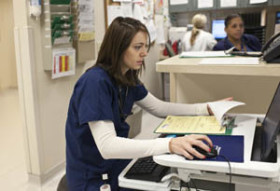 Nurse in scrubs working at a computer station, holding documents and interacting with a computer mouse in a busy hospital setting.
