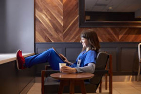 Woman in blue scrubs relaxing in a chair with her feet up, holding a tablet and a cup in a modern lounge area.