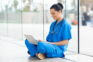 Nurse in blue scrubs sitting on the floor by large windows, working on a laptop and smiling.