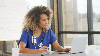 Female healthcare professional in blue scrubs working on a laptop, holding a pen, with a stethoscope around her neck, sitting at a desk in a bright office.