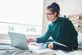 Young woman wearing glasses studying on a bed with a laptop, notebook, and books, in a bright room with natural light from a window.