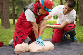 Two people practice CPR on a training mannequin outdoors, with one performing chest compressions and the other providing guidance.