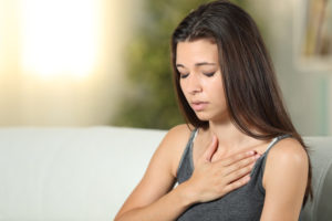 Woman sitting on a couch holding her chest with a concerned expression, possibly experiencing chest pain or discomfort.