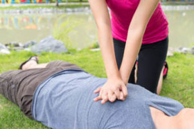 Person performing CPR chest compressions on someone lying on grass outdoors near a pond.