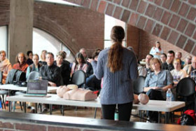 Instructor giving a CPR training session to a seated audience, with CPR mannequins and a laptop on a table in front of the group in a room with brick arches.