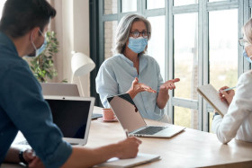 Three people wearing face masks having a discussion around a table with laptops and notepads in a bright, modern office setting.