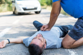 Person performing chest compressions on an unconscious man lying on a road, with a white car in the background.