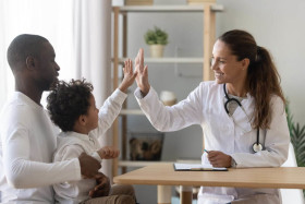 Smiling female doctor giving a high five to a young boy sitting on his father’s lap during a medical consultation in a bright office.