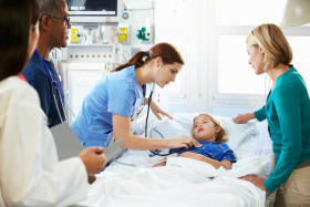 Medical team and a concerned woman attend to a young girl lying in a hospital bed, with a nurse checking the child's vitals using a stethoscope.