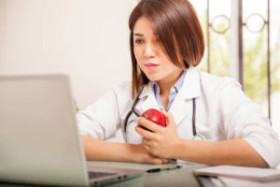 Female doctor in a white coat holding an apple while working on a laptop at her desk.
