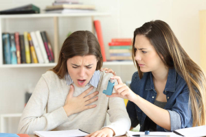 A concerned woman helps another woman having an asthma attack by offering an inhaler indoors, with books and papers on the table in front of them.