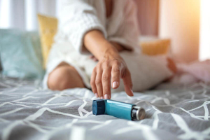 Person in a robe sitting on a bed and reaching for a blue asthma inhaler lying on the patterned bedspread.